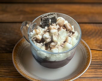 Puede incluir: Una taza de vidrio transparente llena de un postre de chocolate cubierto con crema batida, malvaviscos y una galleta de chocolate. La taza está sobre un platillo blanco con un borde marrón.