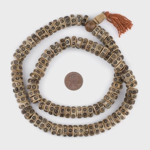May include: A long strand of cylindrical, off-white beads featuring a repeating black circular eye pattern. The strand includes a single larger, round, textured bead and a rust-colored tassel. A copper penny is placed in the center for scale.