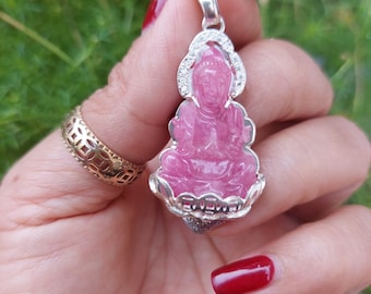 Puede incluir: Un colgante de plata con una piedra tallada de color rosa que representa una figura sentada. El colgante está adornado con pequeñas piedras transparentes alrededor del borde.