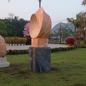 Op de afbeelding: Een modern abstract beeldhouwwerk van oranje en grijs marmer, dat op een sokkel in een grasrijk park staat. Twee rode lantaarns hangen op de achtergrond.
