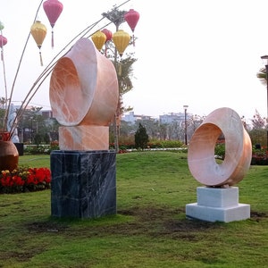 Op de afbeelding: Twee abstracte sculpturen van roze marmer worden tentoongesteld in een park. De sculpturen staan op vierkante sokkels en zijn omgeven door groen gras. De sculptuur links is een ringvorm met een gat in het midden. De sculptuur rechts is een ringvorm met een gat in het midden.