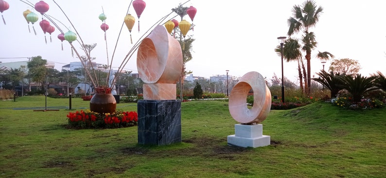 Op de afbeelding: Twee abstracte sculpturen van roze marmer worden tentoongesteld in een park. De sculpturen staan op vierkante sokkels en zijn omgeven door groen gras. De sculptuur links is een ringvorm met een gat in het midden. De sculptuur rechts is een ringvorm met een gat in het midden.