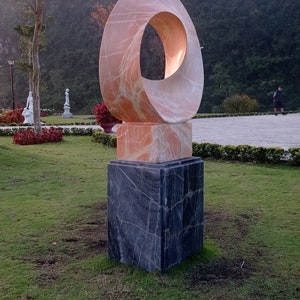 Op de afbeelding: Een roze marmeren sculptuur van een cirkel met een gat in het midden, staande op een zwarte marmeren basis. Het beeld staat in een grasveld met een berg op de achtergrond.