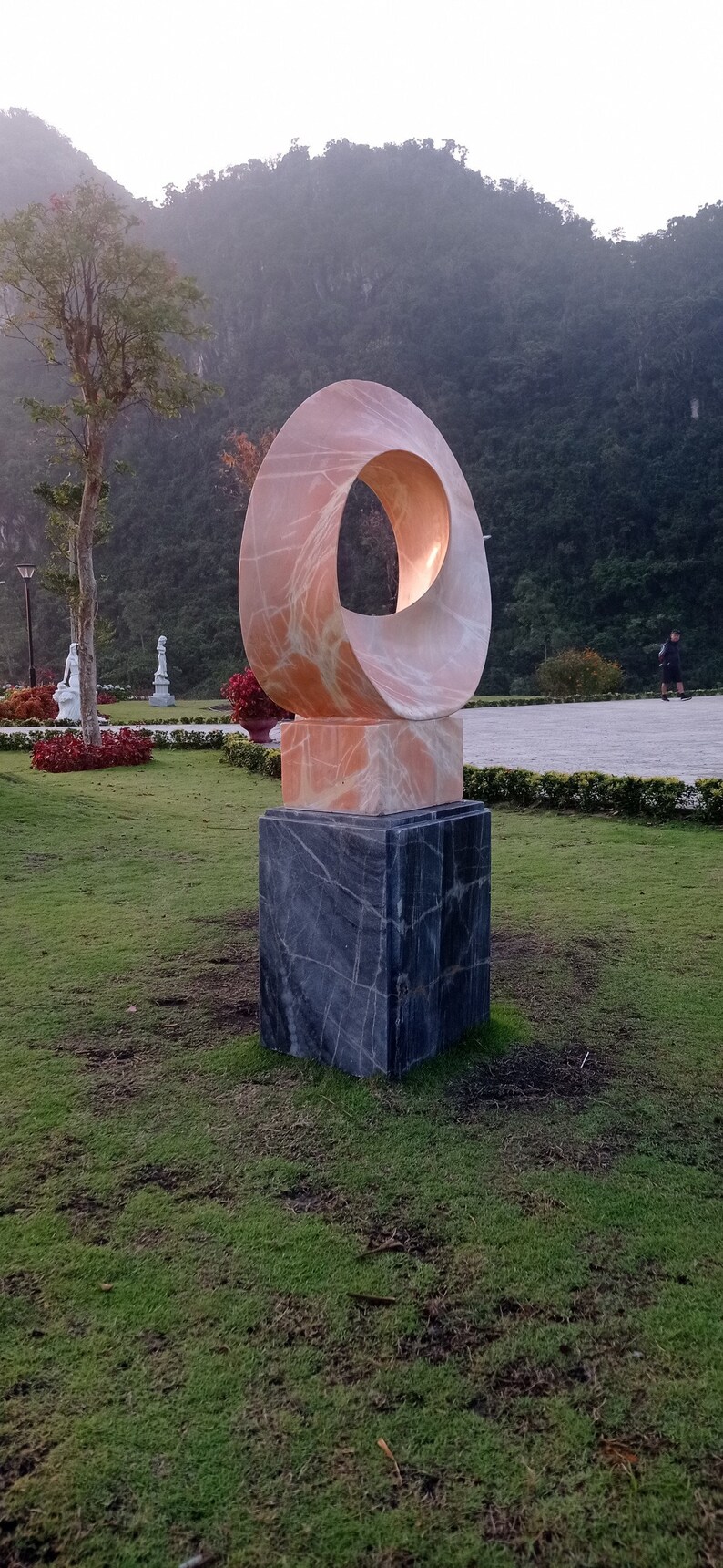 Op de afbeelding: Een roze marmeren sculptuur van een cirkel met een gat in het midden, staande op een zwarte marmeren basis. Het beeld staat in een grasveld met een berg op de achtergrond.