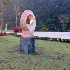 Op de afbeelding: Een roze en witte abstracte sculptuur met een ronde opening in het midden, staande op een zwarte vierkante basis. De sculptuur staat in een park met groen gras en bomen op de achtergrond.