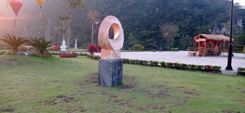 Op de afbeelding: Een roze en witte abstracte sculptuur met een ronde opening in het midden, staande op een zwarte vierkante basis. De sculptuur staat in een park met groen gras en bomen op de achtergrond.