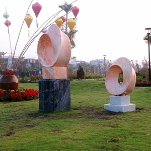 Op de afbeelding: Twee abstracte sculpturen gemaakt van roze en witte steen, met een cirkelvormig ontwerp, worden tentoongesteld in een groene parksetting. De sculpturen staan op sokkels en zijn omringd door groen gras en bloemen.