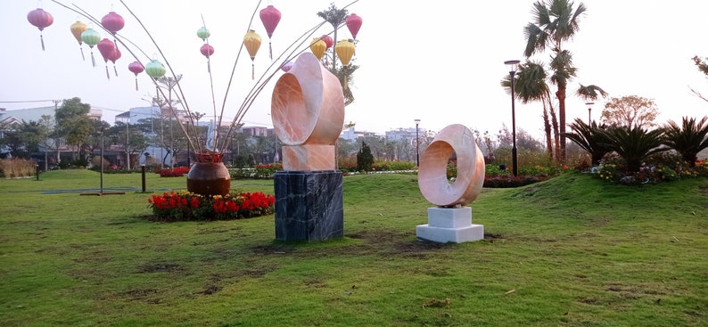 Op de afbeelding: Twee abstracte sculpturen gemaakt van roze en witte steen, met een cirkelvormig ontwerp, worden tentoongesteld in een groene parksetting. De sculpturen staan op sokkels en zijn omringd door groen gras en bloemen.