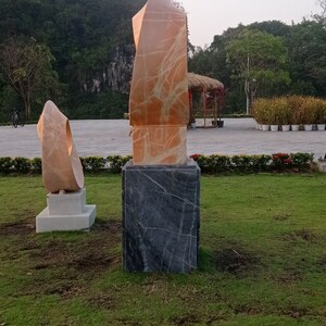 Op de afbeelding: Twee abstracte sculpturen van marmer, een is een hoge, dunne, oranje sculptuur op een zwarte basis, de andere is een kortere, rondere, witte sculptuur op een witte basis. De sculpturen staan in een grasrijk park.