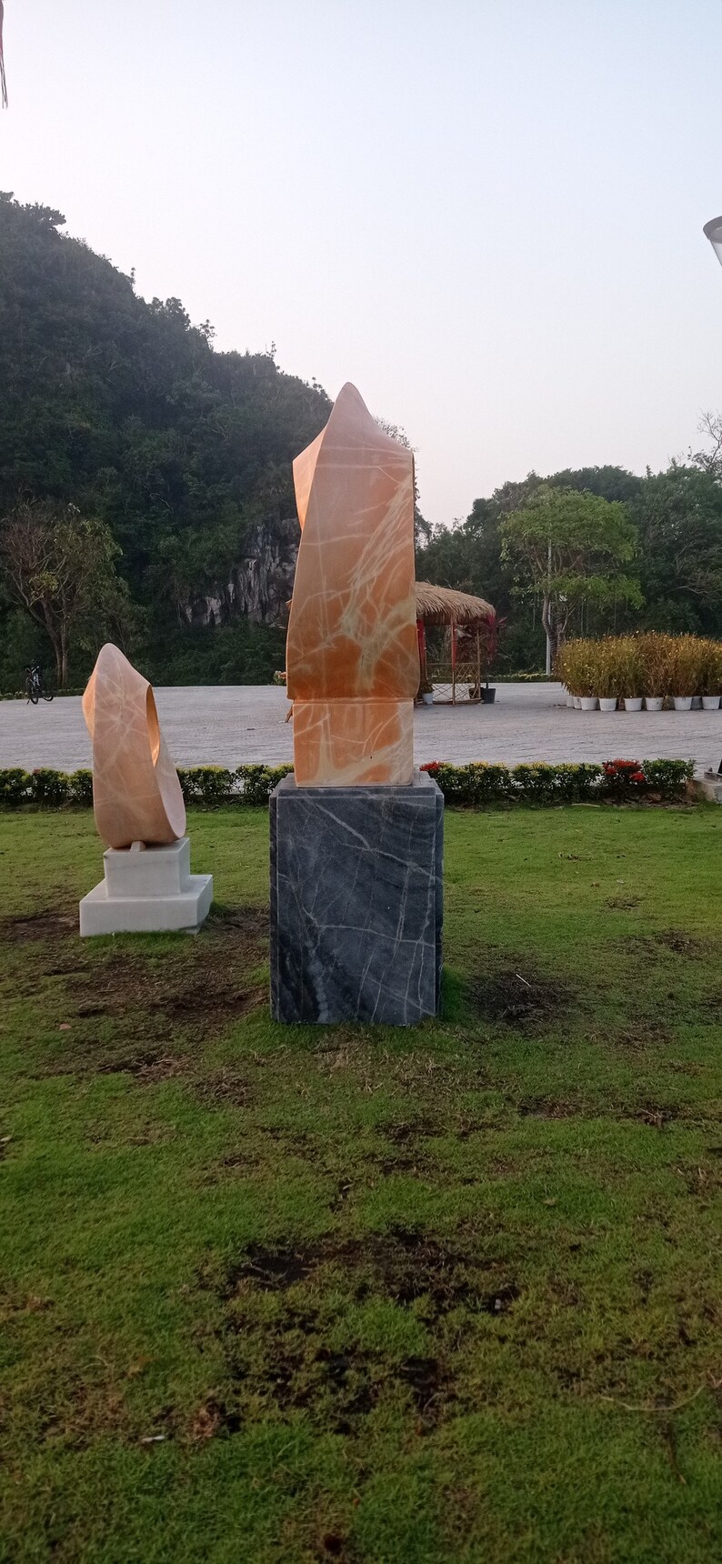 Op de afbeelding: Twee abstracte sculpturen van marmer, een is een hoge, dunne, oranje sculptuur op een zwarte basis, de andere is een kortere, rondere, witte sculptuur op een witte basis. De sculpturen staan in een grasrijk park.