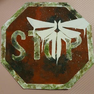 Op de afbeelding: Een handgeschilderd, achthoekig bord met een verweerde uitstraling. Het bord is voornamelijk donkerrood met witte verfspatten. Het woord "STOP" is zichtbaar in het wit, met een wit vuurvliegontwerp. De rand is groen.