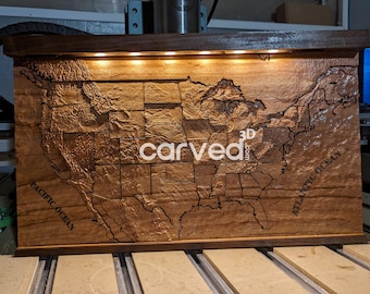 May include: A wooden map of the United States with state borders and the words "PACIFIC OCEAN" and "ATLANTIC OCEAN" carved into the wood. The map is lit from above with LED lights and has a dark wood frame.