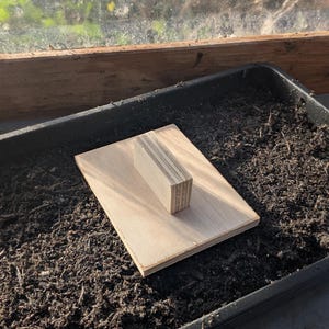 May include: A wooden hand tool, possibly for gardening, sits on a rectangular wooden base. The tool is made of layered wood and rests on dark soil inside a black tray, suggesting a planting or potting setup. The background includes a window.