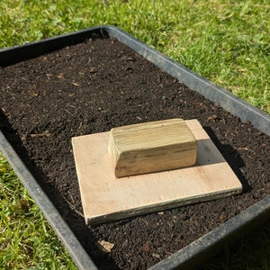 May include: A black plastic seed tray filled with dark soil, a light-coloured wooden board, and a small wooden block. The tray is on green grass, indicating a gardening or planting setup.