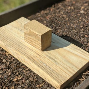 Op de afbeelding: Een houten plank met een kleiner houten blok erop, beide zijn lichtbruin van kleur en hebben een natuurlijke houtnerf. De plank ligt op een bed van donkerbruine mulch.