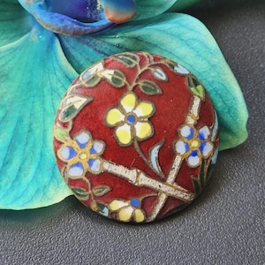 May include: A round cloisonné button featuring a deep red background with a floral pattern. The design includes yellow and blue flowers with white petals, green leaves, and thin gold-toned metal wire outlines separating the colored enamel sections.