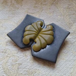 Könnte beinhalten: Eine zweiteilige Brosche mit dunkel anthrazitfarbenen geometrischen Formen und glatten Oberflächen. In der Mitte befindet sich ein helles olivgrünes Dekorelement mit einem symmetrischen, fächerartigen Schnitzmuster, das die dunklen Teile verbindet.