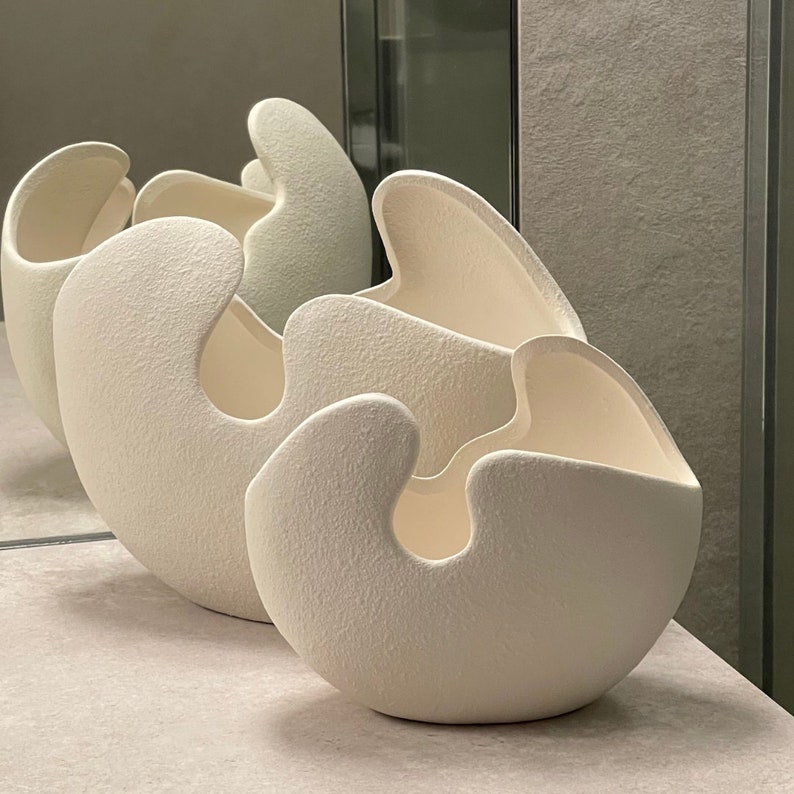 May include: Three off-white, textured ceramic vases with organic, flowing shapes. The vases have a rounded base and a curved opening, creating a unique, modern design. The vases are arranged in a row, with the largest in the foreground.