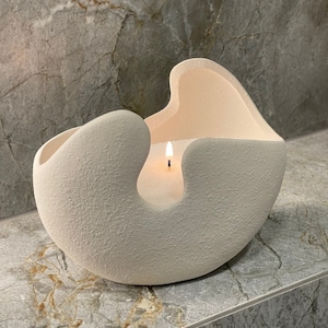 May include: A cream-coloured, textured candle in a unique, organic-shaped holder. The candle is lit, with a small flame visible. The holder has a modern, sculptural design, adding a touch of elegance. A decorative home accent.