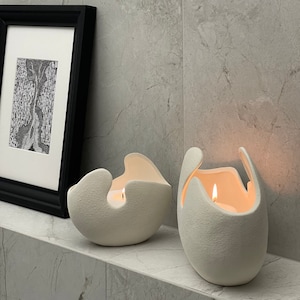 May include: Two off-white, textured ceramic candle holders with lit candles inside. The holders have unique, organic shapes with curved edges. A framed black and white artwork is in the background, adding a touch of artistic flair to the scene.
