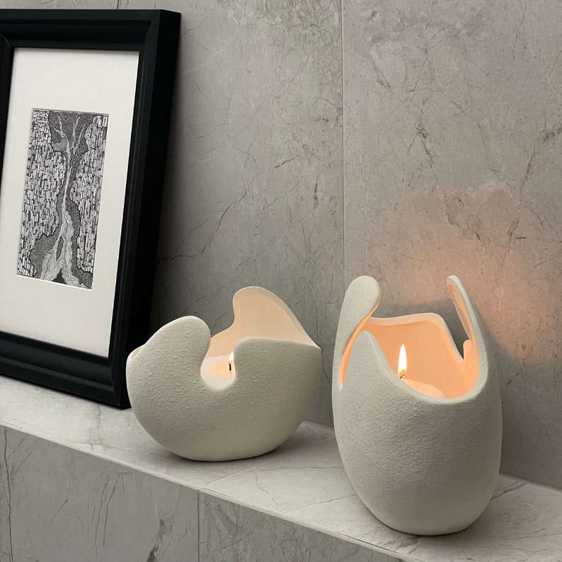 May include: Two off-white, textured ceramic candle holders with lit candles inside. The holders have unique, organic shapes with curved edges. A framed black and white artwork is in the background, adding a touch of artistic flair to the scene.