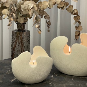 May include: Two off-white, textured, organic-shaped candles with a cutout design. Each candle has a lit wick, casting a warm glow. A dark glass vase with dried eucalyptus branches is in the background, adding a natural element to the scene.