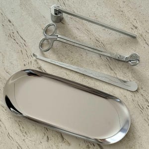 Puede incluir: Un conjunto de accesorios plateados para velas, que incluye un recortador de mecha, un apagador y unas tijeras, colocados cerca de una bandeja ovalada plateada. Las herramientas son de metal brillante y están diseñadas para el cuidado de las velas.