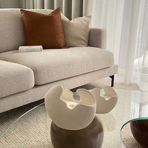 May include: A modern living room scene featuring a beige sofa with brown and beige pillows. Two off-white, organic-shaped candle holders sit on a glass coffee table. A book titled "What Great Paintings Say" is on the sofa.