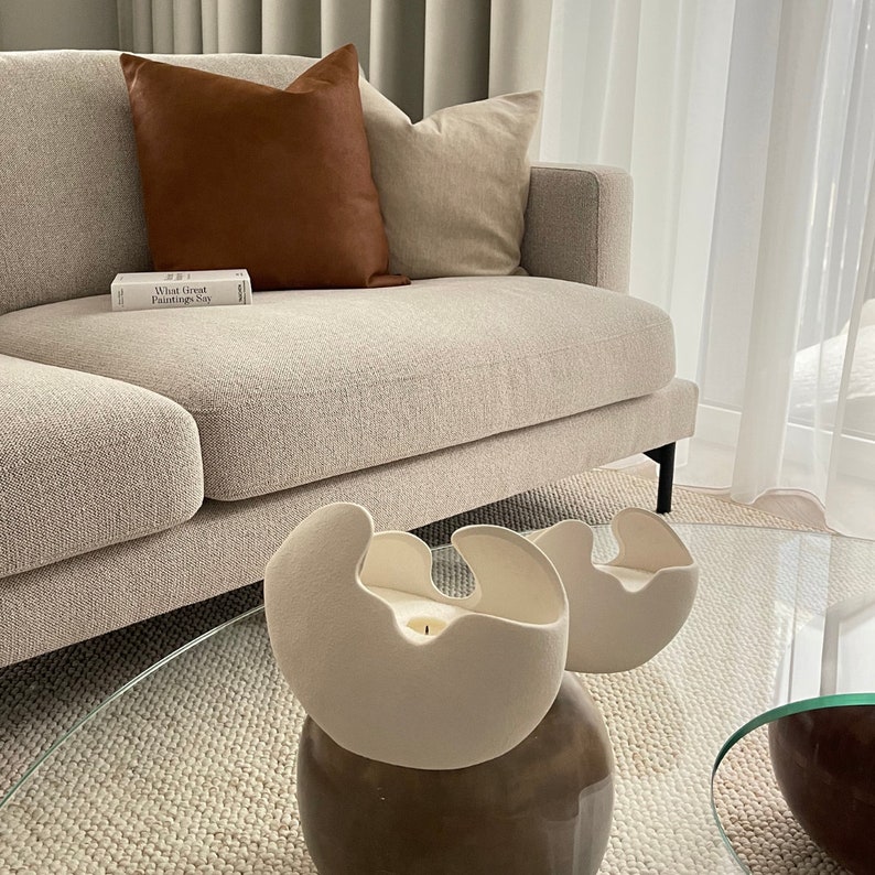 May include: A modern living room scene featuring a beige sofa with brown and beige pillows. Two off-white, organic-shaped candle holders sit on a glass coffee table. A book titled "What Great Paintings Say" is on the sofa.