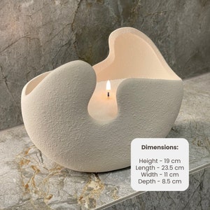 May include: A lit, off-white candle in a textured, abstract-shaped ceramic container. The candle's dimensions are: Height - 19 cm, Length - 23.5 cm, Width - 11 cm, Depth - 8.5 cm. The candle sits on a gray surface.