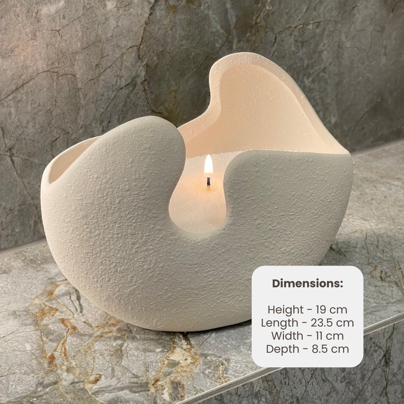 May include: A lit, off-white candle in a textured, abstract-shaped ceramic container. The candle's dimensions are: Height - 19 cm, Length - 23.5 cm, Width - 11 cm, Depth - 8.5 cm. The candle sits on a gray surface.