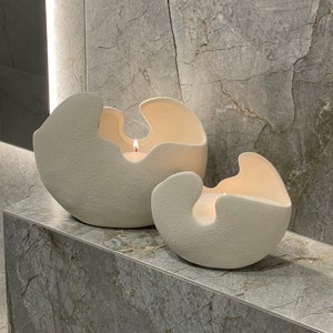 May include: Two off-white, textured ceramic candle holders with a unique, organic shape. Each holder has a large opening and contains a lit candle, casting a warm glow. The holders are displayed on a marble surface.