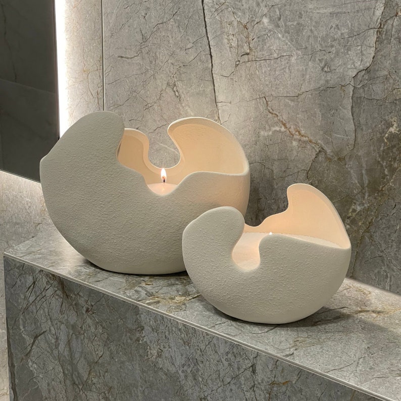 May include: Two off-white, textured ceramic candle holders with a unique, organic shape. Each holder has a large opening and contains a lit candle, casting a warm glow. The holders are displayed on a marble surface.