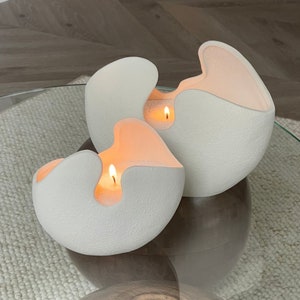 May include: Two white, textured, sculptural candles with a unique design. The candles have an organic shape with a cutout revealing the glowing, orange-toned wax and lit wicks. The candles are on a glass table.
