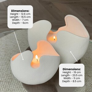 Op de afbeelding: Drie witte, getextureerde, eivormige kaarsen met een uniek ontwerp. Elke kaars heeft een uitsparing die een warme, oranje gloed van de brandende lont onthult. De grotere kaars is 19 cm hoog, 23,5 cm lang, 11 cm breed en 8,5 cm diep.