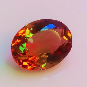 Puede incluir: Una piedra preciosa grande de forma ovalada con un brillo iridiscente rojo, naranja y verde. La piedra preciosa está tallada con muchas facetas y tiene un alto pulido.