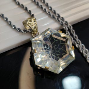 May include: A clear, hexagonal pendant necklace with a detailed, geometric design. The pendant is suspended from a silver-colored rope chain. The pendant's setting is gold-toned, and the bail has an ornate, openwork design.