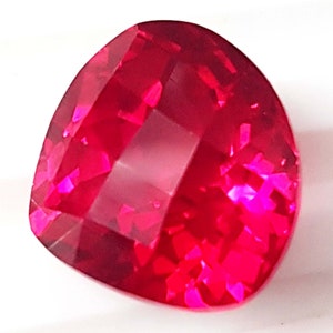 May include: A deep red gemstone with a cushion cut and a faceted finish. The gemstone is set in a clear, transparent setting.