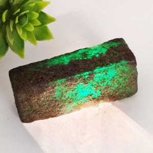 Könnte beinhalten: Ein rauer, dunkelgrüner Smaragd mit einem leuchtend grünen Schimmer. Der Stein hat eine rechteckige Form und eine strukturierte Oberfläche.