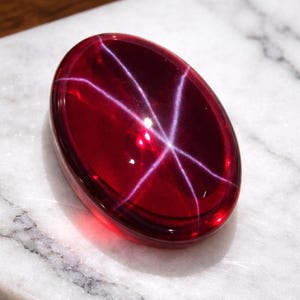 Könnte beinhalten: Ein polierter, oval geformter, tiefroter Edelstein mit einem sechsstrahligen Sternmuster. Der Edelstein liegt auf einer weißen Marmoroberfläche, die seine satte Farbe und seinen einzigartigen optischen Effekt hervorhebt. Das Sternmuster entsteht durch Lichtreflexionen.