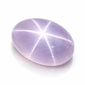 Pode incluir: Uma pedra preciosa ovalada, de cor lavanda, com um padrão de estrela de seis raios. O efeito de estrela é criado pela luz que reflete nas inclusões dentro da pedra. A gema está sobre um fundo branco.
