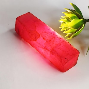 May include: A vibrant pink, rectangular, rough-cut gemstone. The gemstone has a translucent appearance and is illuminated from the side, highlighting its natural texture.