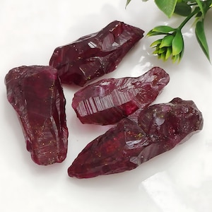 Op de afbeelding: Vier ruwe robijnkristallen, elk met een dieprode kleur en een puntige vorm. De kristallen hebben een natuurlijke, ruwe textuur en zijn op een wit oppervlak gerangschikt.