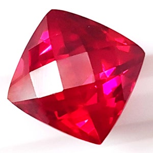 May include: A deep red gemstone with a square cushion cut. The gemstone has a high polish and is faceted.
