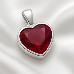 May include: A heart-shaped pendant featuring a faceted, deep red gemstone set in a silver-coloured metal. The pendant has a loop at the top for a chain. The gemstone is the focal point, reflecting its rich colour and catching the light.