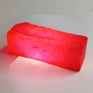 Może przedstawiać: Duży, prostokątny, jaskrawoczerwony rubin. Rubin jest surowy i nieoszlifowany.