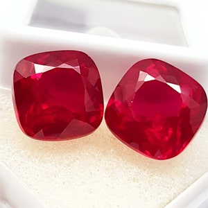 Puede incluir: Dos rubíes tallados en cojín, facetados. Las piedras preciosas son de un color rojo intenso y vibrante y se muestran en una caja blanca. Las facetas captan la luz, creando un efecto brillante. Estos rubíes son probablemente para la fabricación de joyas.