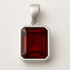May include: A rectangular pendant with a deep red gemstone set in a silver-colored metal frame. The gemstone has a faceted cut, and the pendant has a loop for a chain.