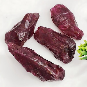 Może przedstawiać: Cztery surowe kryształy rubinu, każdy o głębokim czerwonym kolorze i spiczastym, fasetowanym kształcie. Kryształy są ułożone na białym tle.