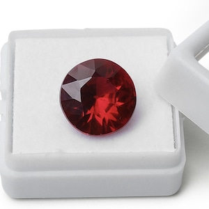 May include: A deep red gemstone with a round cut, displayed in a white box.